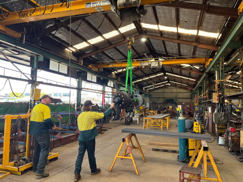 Workers in high-vis clothing using an overhead crane to lift a heavy industrial component inside a workshop Workers in high-vis clothing using an overhead crane to lift a heavy industrial component inside a workshop