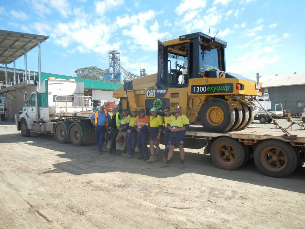A group of workers standing next to a low-loader carrying a Caterpillar compactor, representing teamwork in machinery logistics