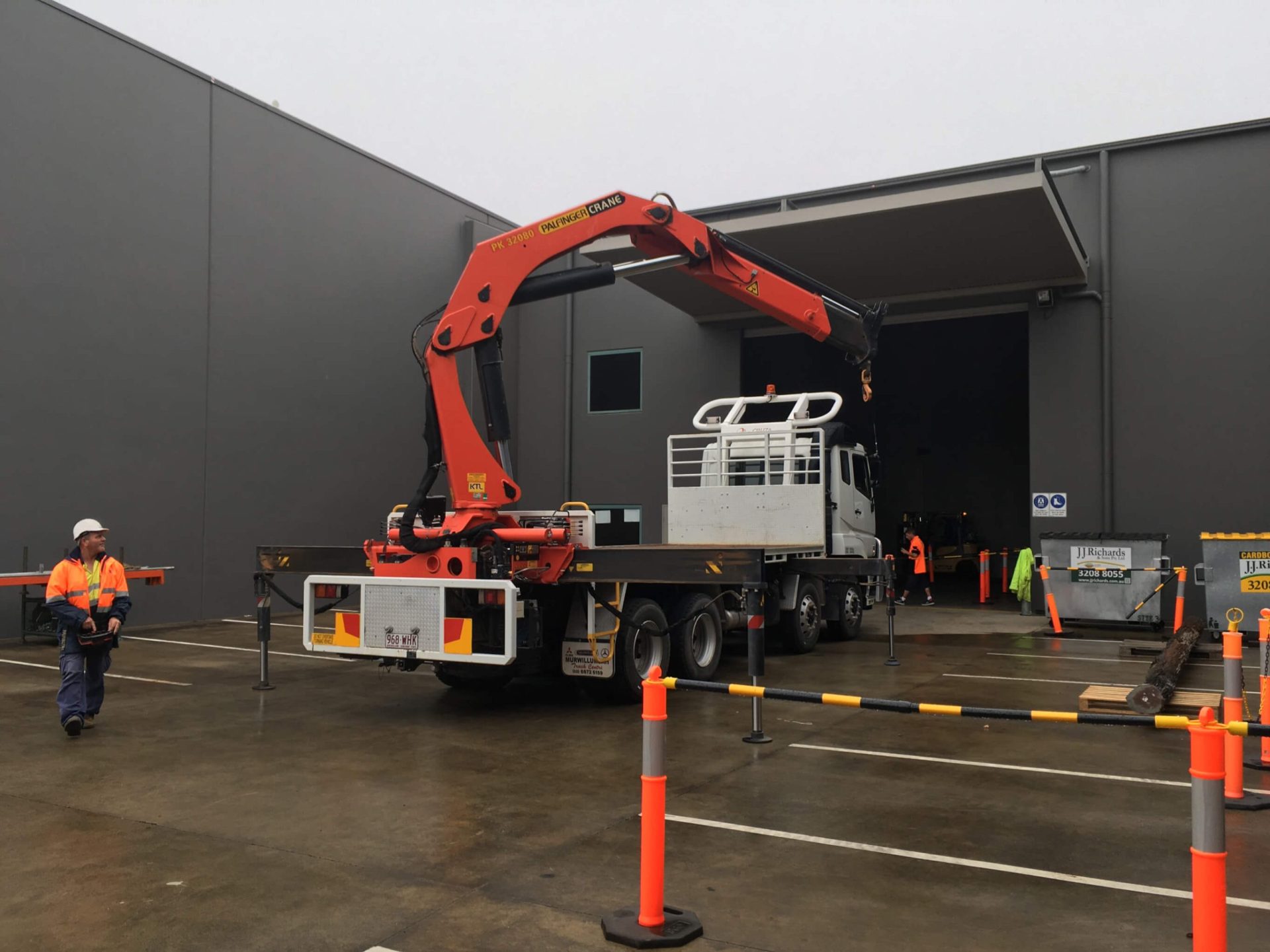 A truck-mounted Palfinger crane lifting materials outside an industrial facility, with workers in high-vis gear ensuring safety